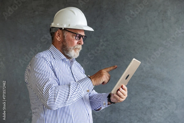Fototapeta An older engineer wearing a white safety helmet uses a digital tablet, symbolizing modern construction practices, technology integration, and innovation in engineering.