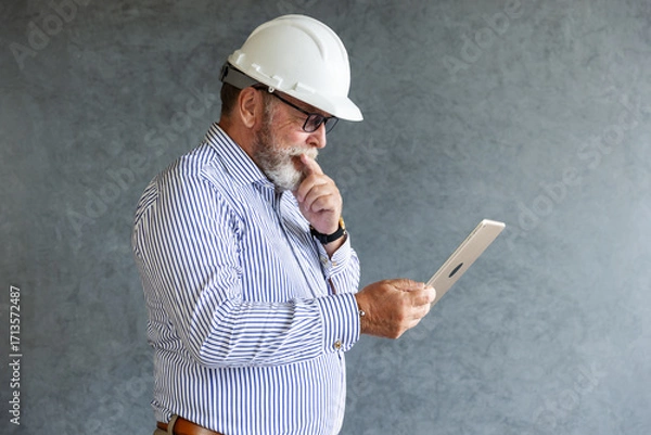 Fototapeta An older engineer wearing a white safety helmet uses a digital tablet, symbolizing modern construction practices, technology integration, and innovation in engineering.