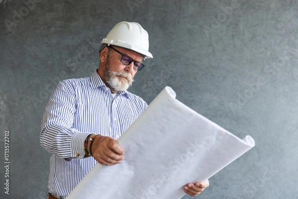 Fototapeta An experienced male engineer wearing a white safety helmet studies construction blueprints, symbolizing architecture, planning, and expertise in building and civil engineering projects.