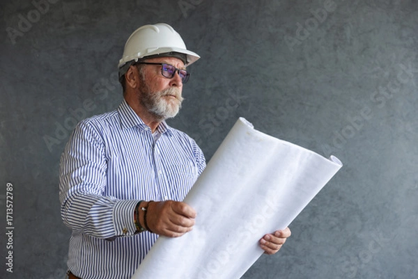 Fototapeta An experienced male engineer wearing a white safety helmet studies construction blueprints, symbolizing architecture, planning, and expertise in building and civil engineering projects.