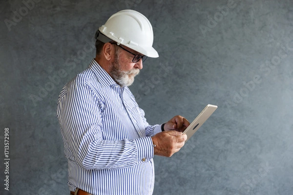 Fototapeta An older engineer wearing a white safety helmet uses a digital tablet, symbolizing modern construction practices, technology integration, and innovation in engineering.