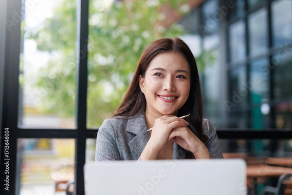 Fototapeta Smiling Asian businesswoman working on a laptop in a stylish cafe, enjoying a productive moment filled with natural light and greenery