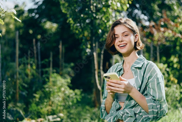 Fototapeta Portrait of a cheerful woman outdoors, smiling while looking at her phone, surrounded by lush trees and vibrant greenery.