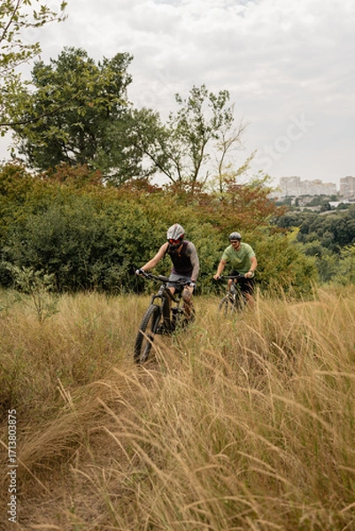 Fototapeta Cyclists enjoy a grassy nature path surrounded by trees and plants during daytime.