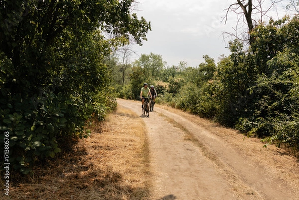 Fototapeta Two people riding bicycles through a dirt path surrounded by lush greenery