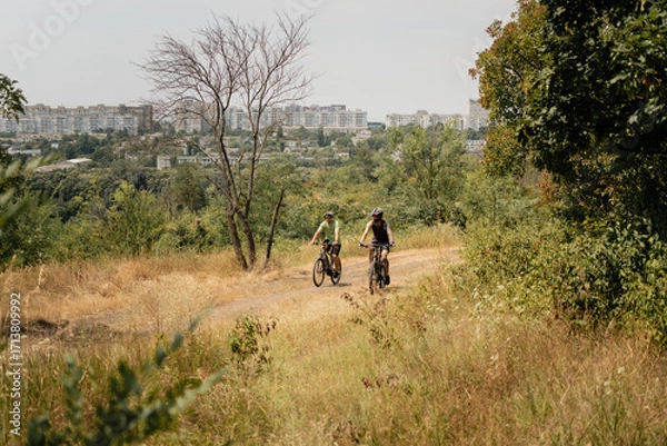 Fototapeta Two people riding bicycles on a dirt trail surrounded by trees and city view