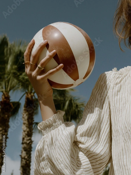 Fototapeta Person holding a beach ball in their hand. the person is wearing a white blouse with a striped pattern and has blonde hair. the beach ball is round and has a brown and white striped pattern on it.