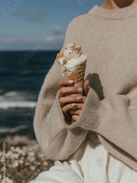 Fototapeta Person holding an ice cream cone in their hand. the person is wearing a beige sweater and is standing on a rocky beach with the ocean in the background.