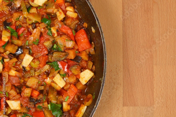 Fototapeta A top-down close-up view of a rustic Italian caponata, a delicious stew of eggplant and other vegetables in a pan, with a wooden background and copy space