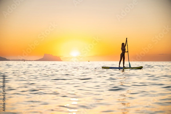 Fototapeta Young woman stand-up paddleboarding at ocean during sunset