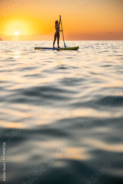 Fototapeta Serene young woman paddleboarding at ocean during sunrise