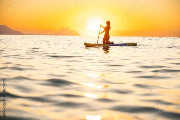 Fototapeta Young woman paddleboarding at ocean during serene sunset