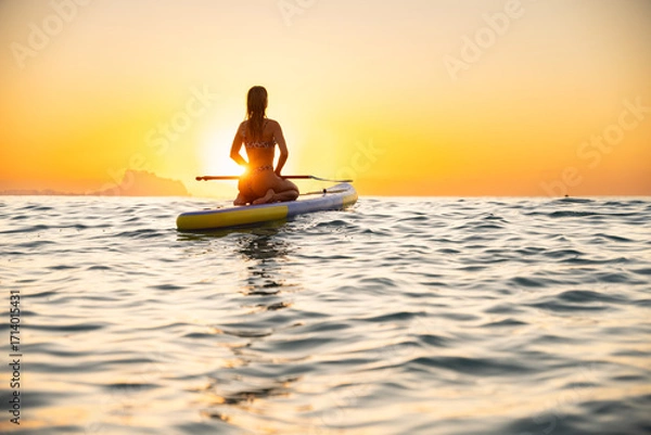 Fototapeta Young woman paddleboarding in serene ocean at sunset
