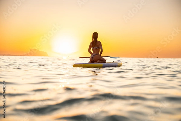 Fototapeta Young woman sitting on paddleboard in calm ocean at sunrise