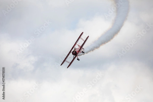 Fototapeta A restored World War II era red biplane airplane performing aerobatic maneuvers in the sky during an air show event, leaving a trail of smoke against a cloudy background in Hillsboro, Oregon, USA