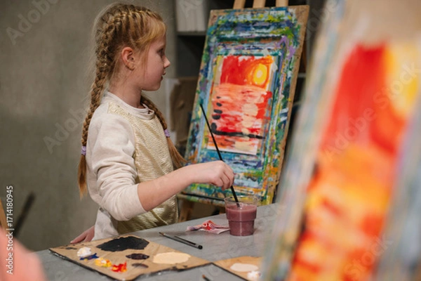 Fototapeta Girl enjoys painting in workshop. Art lesson. Education and learning concept. Child draw a picture in art class. School kid drawing with brush landscapes on easel. Mix and combine colors.
