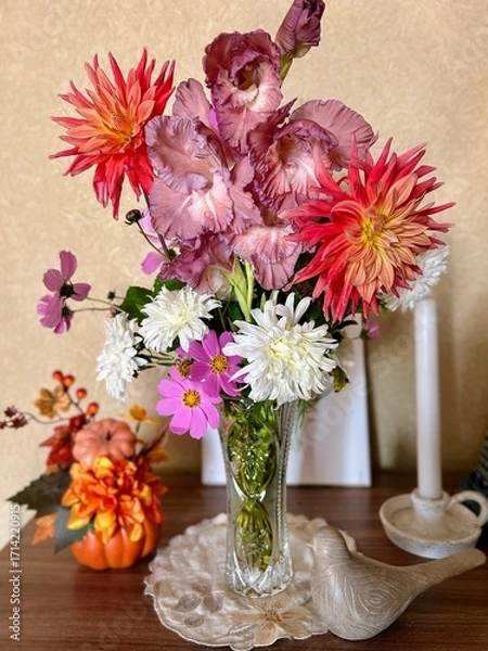 Obraz A colorful bouquet featuring dahlias, gladiolus, and chrysanthemums in a glass vase. Autumn decorations include a small pumpkin and textured bird figurine on a wooden surface.
