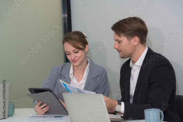 Fototapeta Professional colleagues discussing project documents in modern office setting. woman is reviewing tablet while man is presenting printed materials. Their expressions convey collaboration and focus