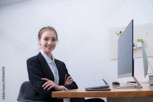 Fototapeta Confident young woman in professional suit sits at modern office desk, smiling warmly. workspace features computer monitor, stationery, and minimalist design, creating productive atmosphere