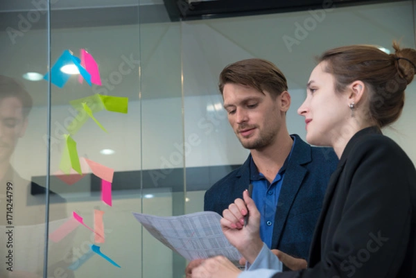 Fototapeta Creative professionals collaborate in modern office setting, discussing ideas while reviewing documents. Colorful sticky notes on glass wall enhance innovative atmosphere