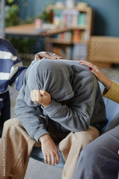 Fototapeta Teenager boy with hood covering head sitting with face in hands, receiving comfort from multiethnic group with supportive hands on shoulder and back, emotional support concept