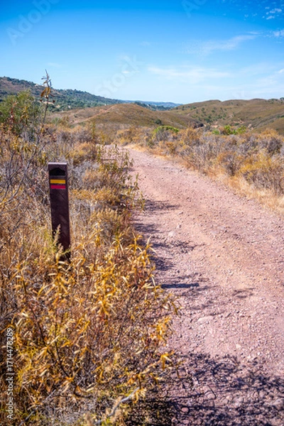 Fototapeta Trail marker on scenic Algarve Portugal hiking trail Via Algarviana dirt path with lush fields rolling hills background