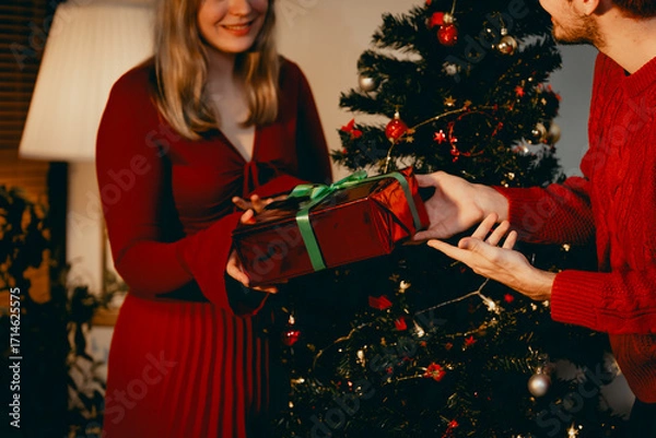 Fototapeta A cheerful group of diverse friends gathers around a decorated Christmas tree, exchanging gifts with laughter and joy at a festive holiday party.