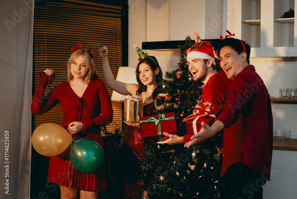 Fototapeta A cheerful group of diverse friends gathers around a decorated Christmas tree, exchanging gifts with laughter and joy at a festive holiday party.