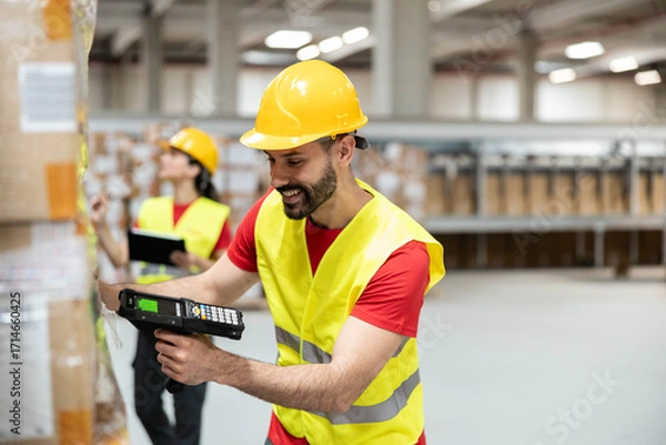 Obraz A cheerful worker scans boxes in a warehouse using a handheld device, showcasing efficiency and teamwork in logistics.