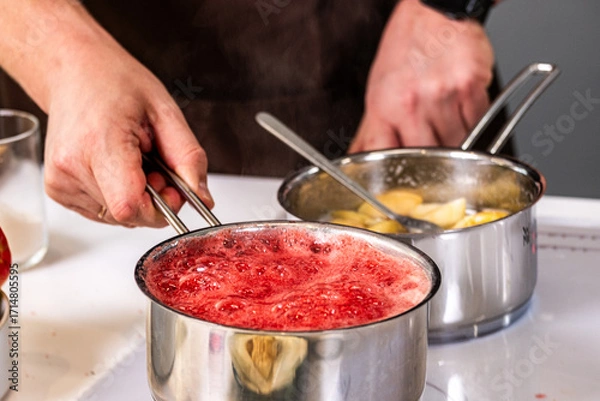 Fototapeta Hands of a chef holding a stainless steel pot with bubbling red sauce, while another pot with pasta simmers on the stove, showcasing culinary skills and vibrant cooking process