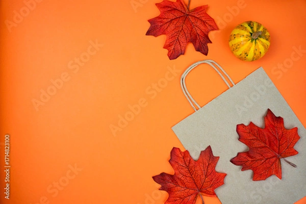 Fototapeta Autumn flatlay composition with paper shopping bag, autumn maple leaves and small pumpkin on an orange background. Fall shopping. Thanksgiving sales concept. Top view. composition. Copy space.