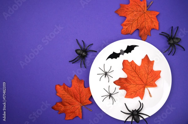 Fototapeta Halloween table setting with a white plate, orange autumn leaves, black spiders and bats on a purple background. Festive and spooky decoration concept for Halloween parties. Top view. Flatlay.