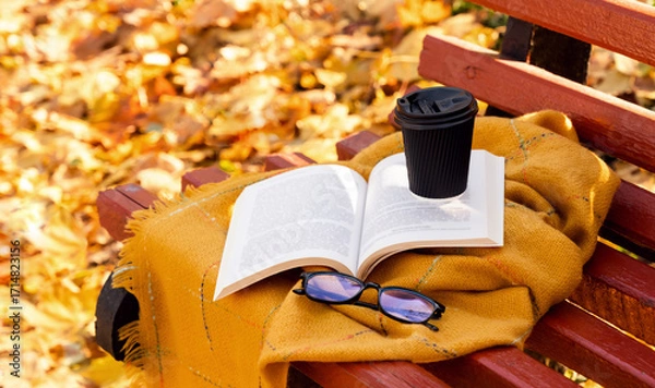 Fototapeta Open book, hot coffee and eyeglasses on a warm blanket outdoors on a bench in autumn park. Cozy fall atmosphere. Reading, studying, and relaxation concept. Selective focus.