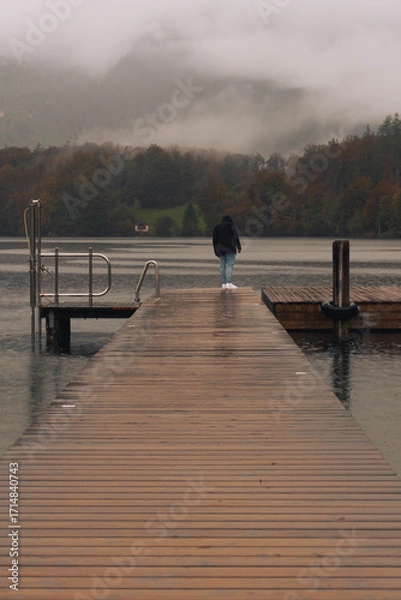Obraz A man walks along the edge of the wooden pier, observing the autumn landscape on a rainy day