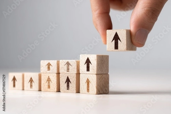 Fototapeta Human hand building a staircase of wooden blocks with arrows pointing upwards showing financial or personal growth