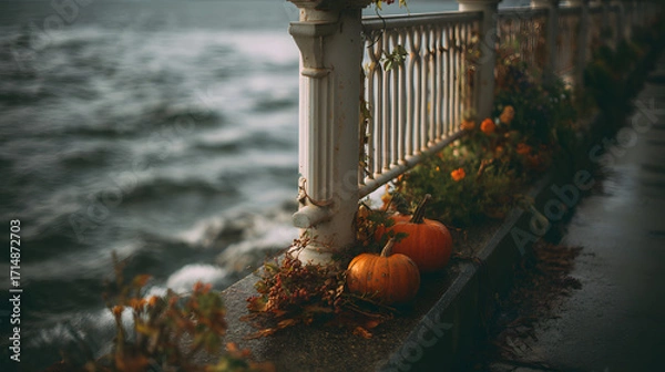 Fototapeta Two orange pumpkins sit beside a waterfront railing, surrounded by autumn foliage and gentle waves, evoking a cozy fall atmosphere.
