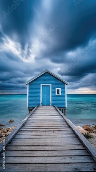 Obraz A serene coastal scene showcases a vibrant blue beach hut atop a wooden pier extending into a tranquil turquoise sea under dramatic skies.