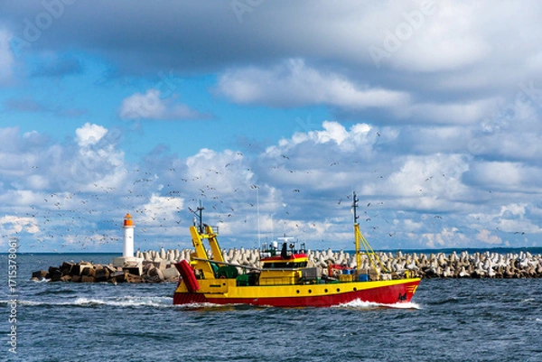 Fototapeta Fishing boat returns home from sea to harbor surrounded by seagulls, with lighthouse in background under dramatic sky, perfect for travel, maritime, fishing industry and coastal projects