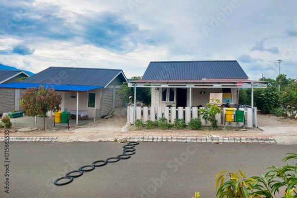 Fototapeta This image shows a row of small permanent houses in a housing complex, featuring blue and black metal roofs, small front yards, and colorful trash bins in front