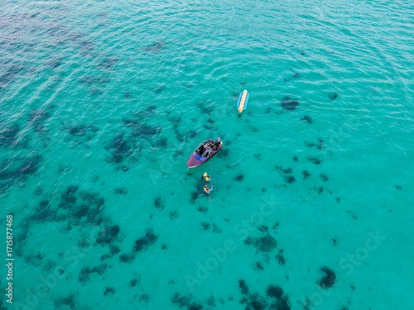 Fototapeta Scenic aerial view of a motorboat and a banana boat resting on calm blue-green waters while tourists with life vests enjoy swimming beside the boat.