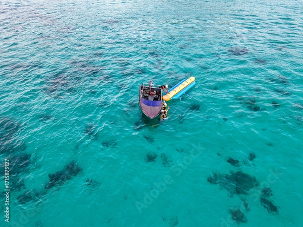 Fototapeta Turquoise ocean scene featuring a small boat and banana boat with several tourists floating over coral formations, captured beautifully from above.