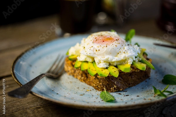 Fototapeta Black bread toast with fried benedict egg, avocado and salmon spread in blue plate with coffee in background, wooden shabby table with coffee pot, glass and books stack