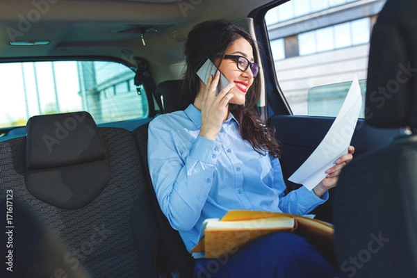 Fototapeta Beautiful young business woman sitting in the car looking at her planner and talking at phone.
