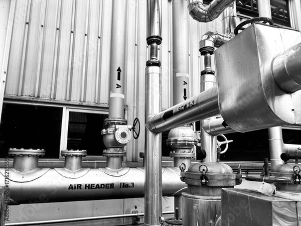 Fototapeta black-and-white image of complex industrial piping and machinery. The detailed composition of valves, tubes, and a corrugated metal wall highlights the intricate and functional aesthetics