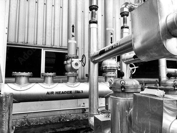 Fototapeta black-and-white image of complex industrial piping and machinery. The detailed composition of valves, tubes, and a corrugated metal wall highlights the intricate and functional aesthetics