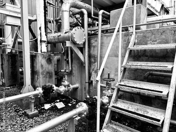 Fototapeta A black and white image of industrial machinery, pipes, and a metal staircase in a gritty factory environment. The photo captures the worn textures and complex layers of an aging facility, conveying 
