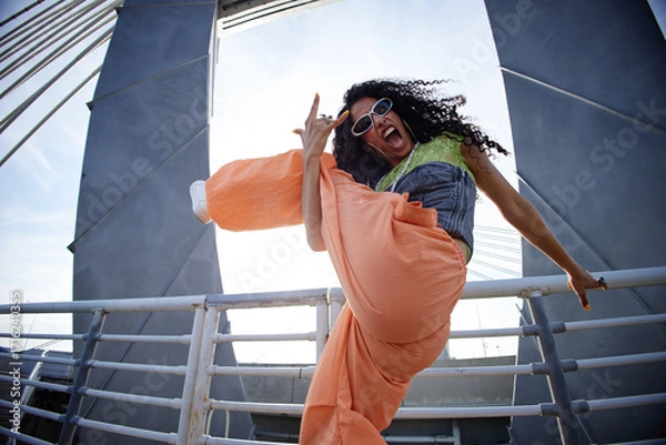 Fototapeta Female zoomer with long curly hair wearing sunglasses energetically kicking leg up and making hand gesture while standing on urban bridge, expressing excitement and confidence