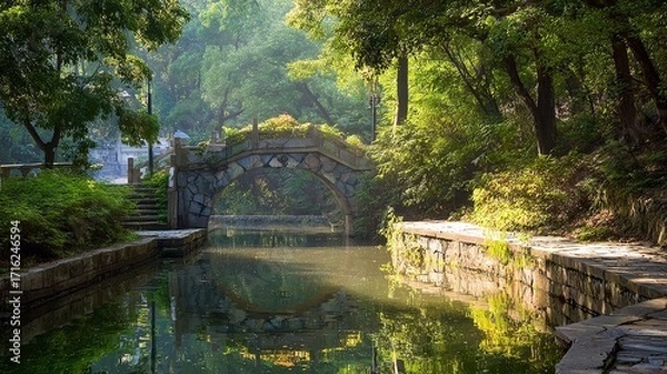 Fototapeta Stone arch bridge over calm water in a lush green park with reflections and trees