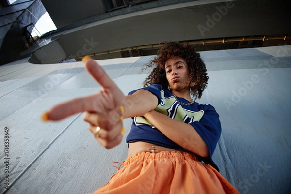 Fototapeta Woman standing outdoors crossing arms and pointing finger toward camera with confident expression, curly hair visible, urban setting in background, low angle view