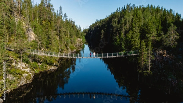 Fototapeta Visitors walk across a hanging suspension bridge over the serene Canyon Lake Julma Olkky, surrounded by lush forests in Hossa National Park in Finland